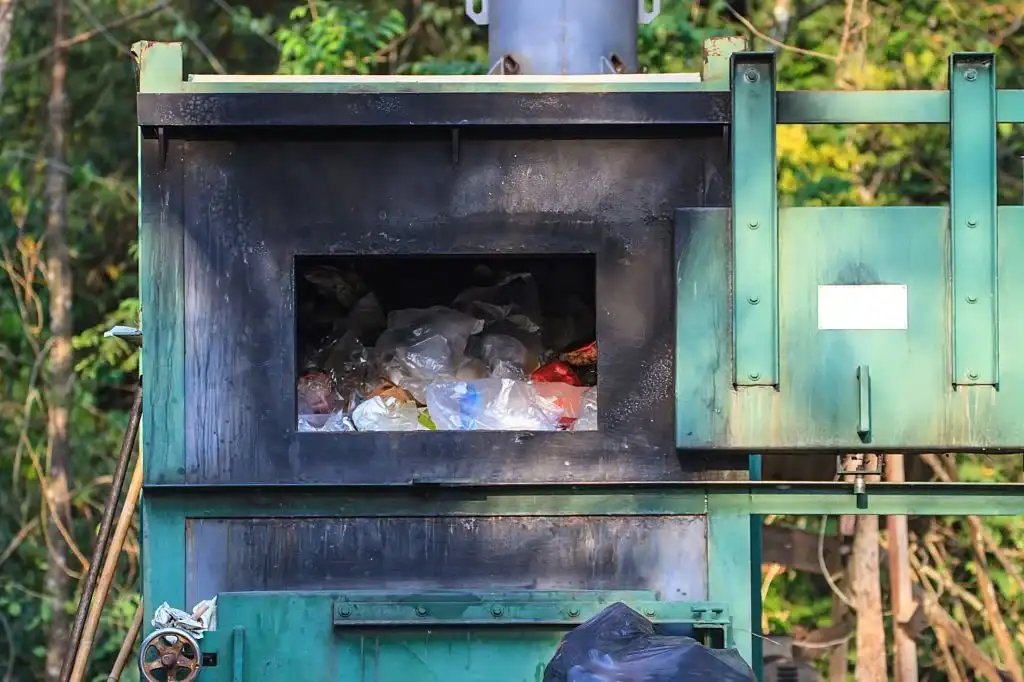 A green, industrial waste incinerator filled with trash, set against a natural backdrop.