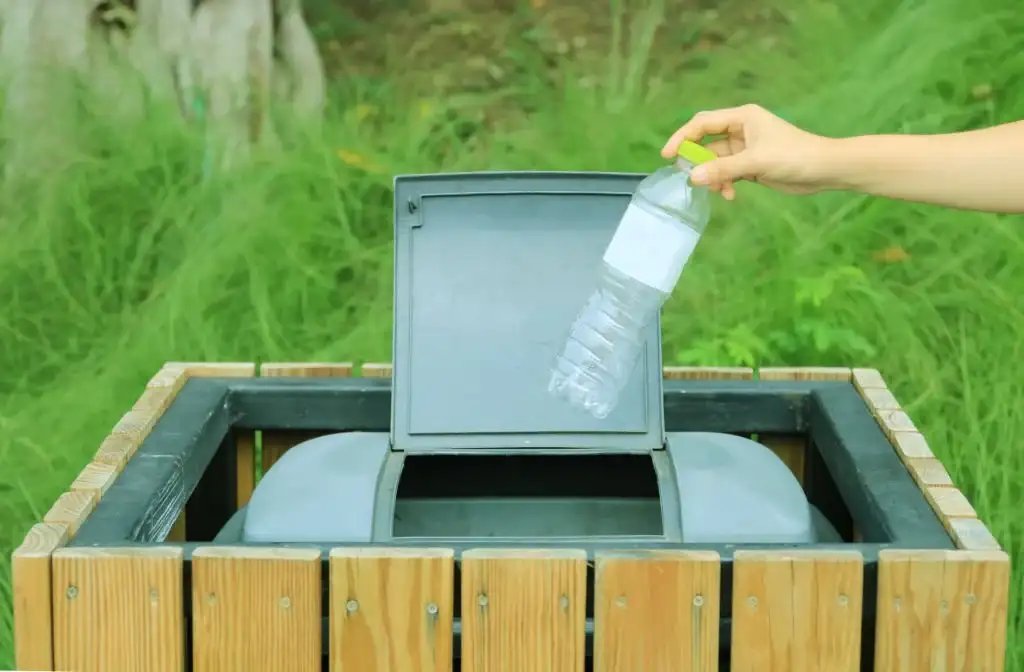 A hand disposing of a plastic bottle into a recycling bin in a grassy area.
