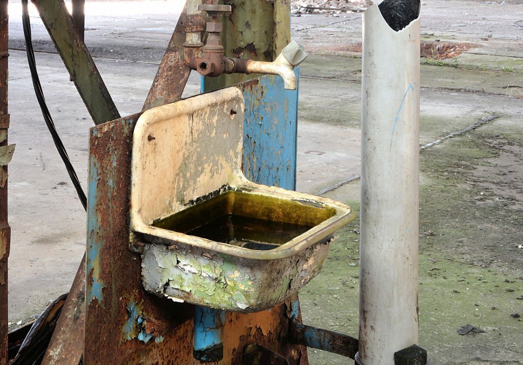 An old, rusted industrial sink with peeling blue and yellow paint in a dilapidated factory setting.