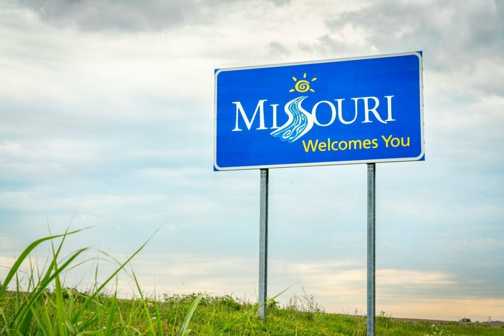A blue "Missouri Welcomes You" sign against a cloudy sky with green grass below.