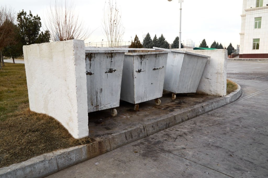 Three large, weathered outdoor trash bins grouped together on a concrete surface.